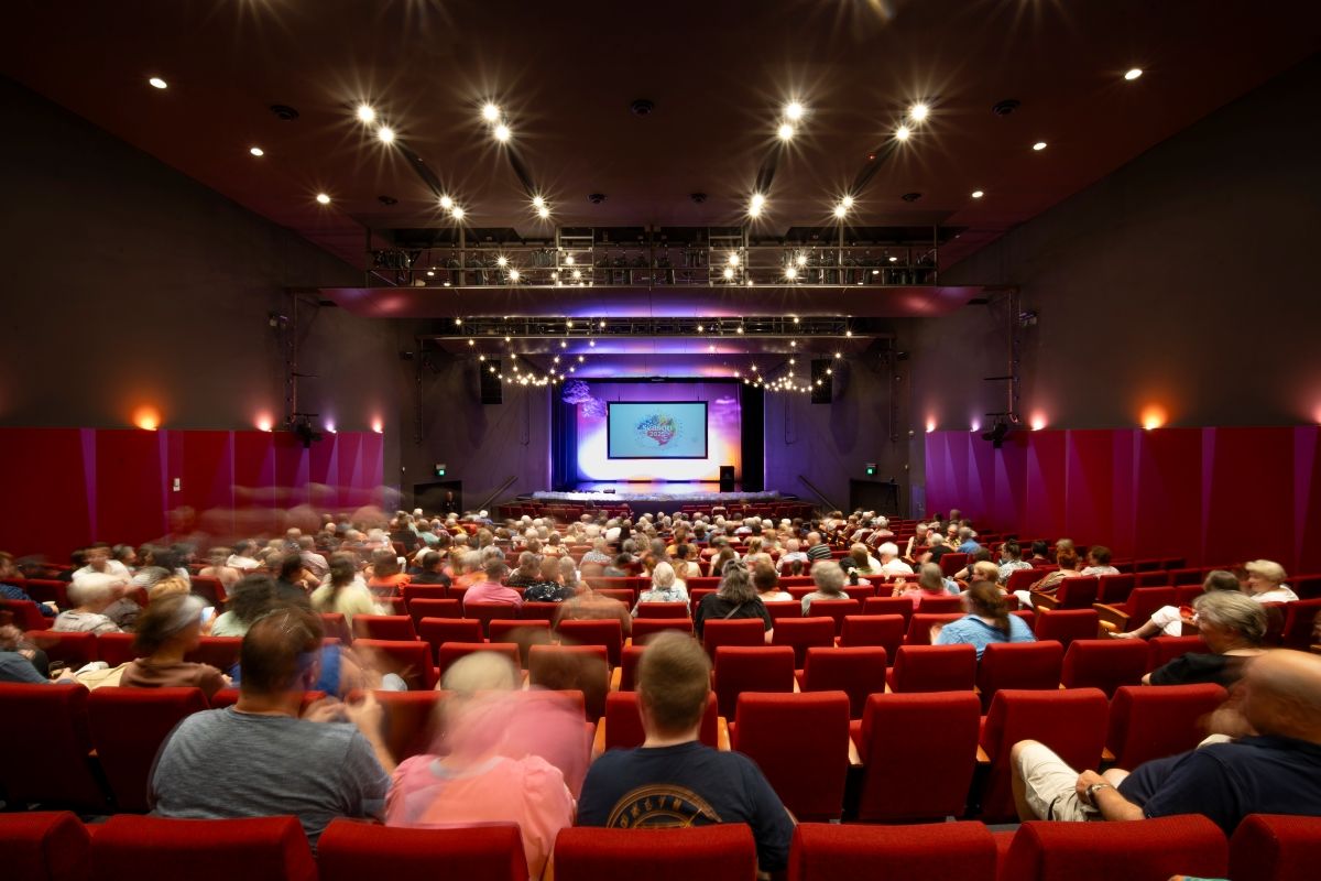 A large crowd of people sitting inside a theatre auditorium. The stage is lit.