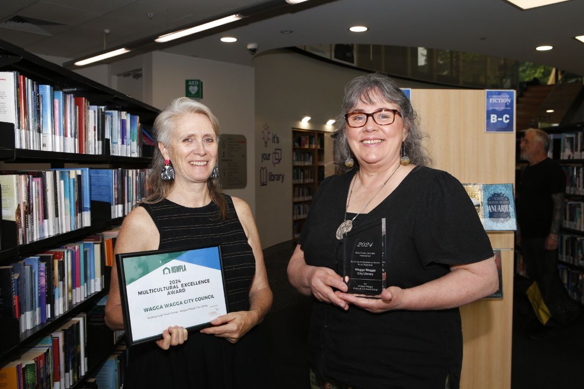 Two women smiling to camera and holding awards and standing between aisles of bookshelves at a library.