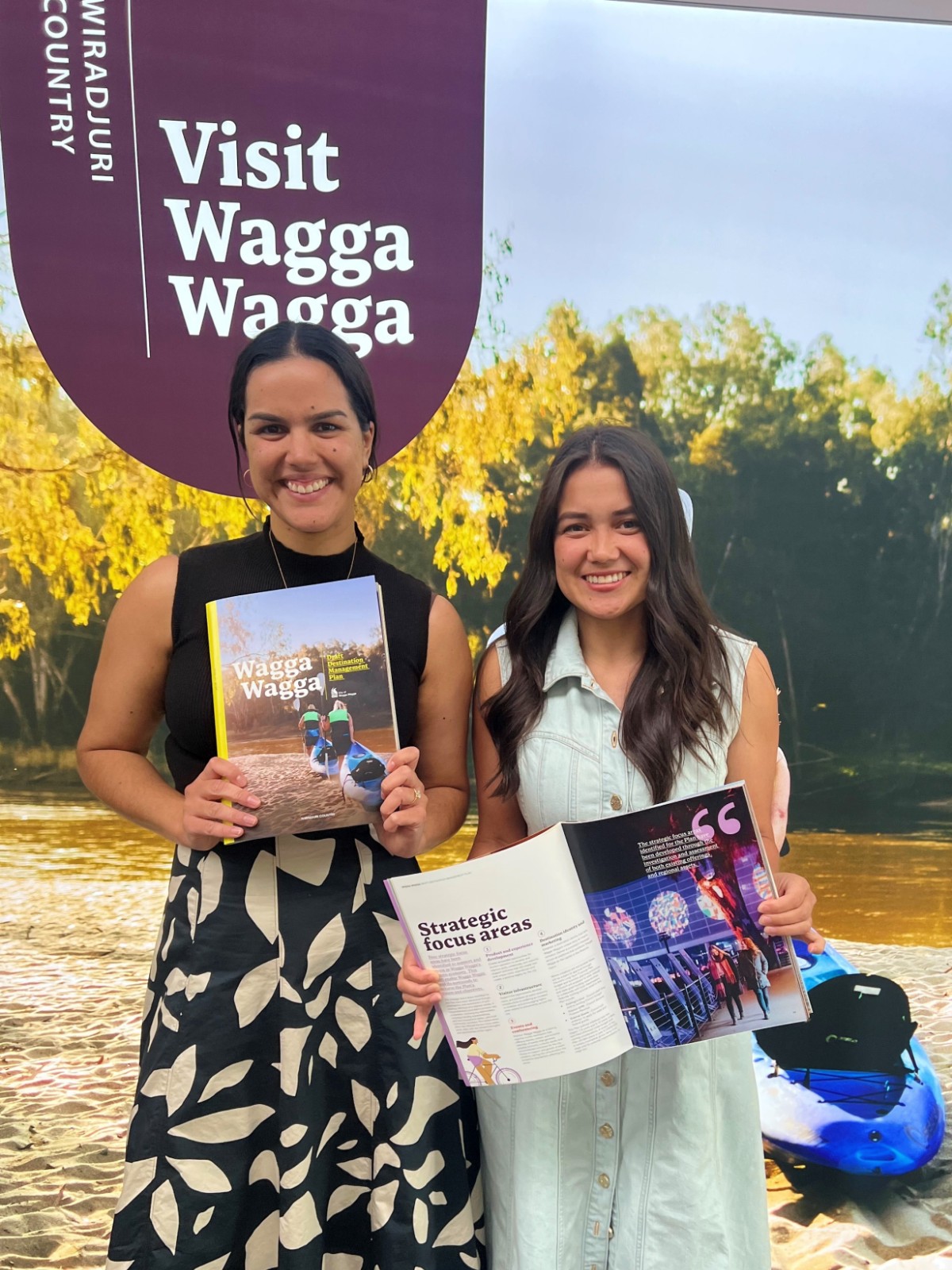 Two women holding prnted copies of the Destination Management Plan, while standing in front of a pull-up promotion sign for the plan.