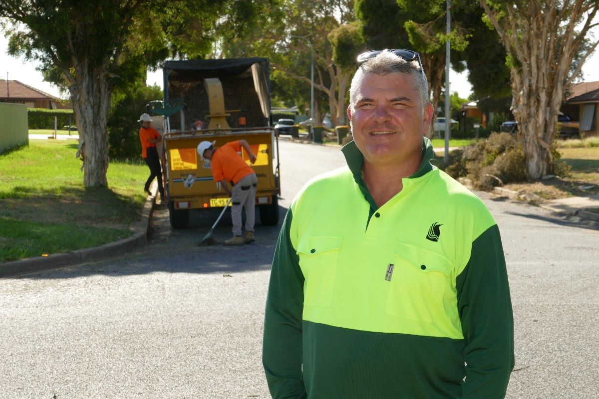 Man in hi-vis shirt standing beside a suburban road with a wood chipping truck in the background.