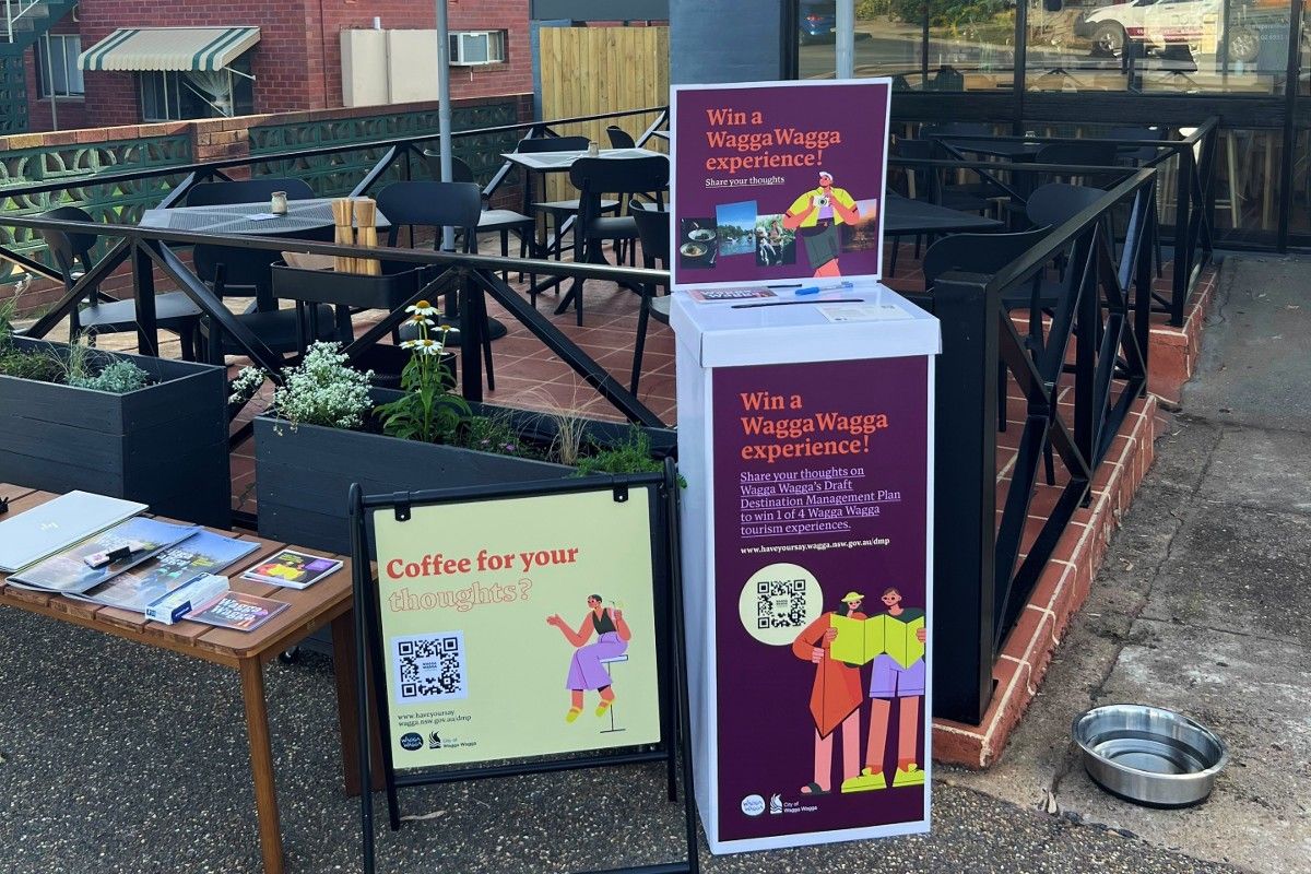 : Information pop-up stall at a café, with small table with information flyers and booklets sitting on it, a sign saying coffee for your thoughts, and an upright box for entry forms.