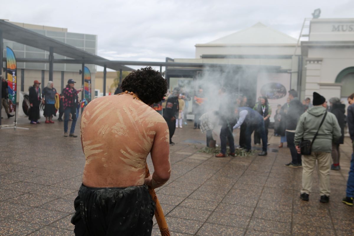 A young Aboriginal man wearing cloth and pigment on his skin plays a Didgeridoo while people walk through the smoke of a smoking ceremony.