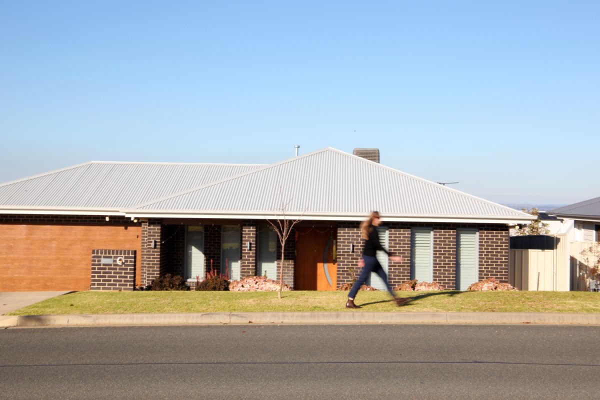 A woman walks briskly past a modern single-storey brick house on a sunny day. The house has a dark brick exterior, a sloped metal roof, and a wooden front door. The sky is clear and blue, and the street in front is quiet and well-maintained. The woman appears slightly blurred in motion.