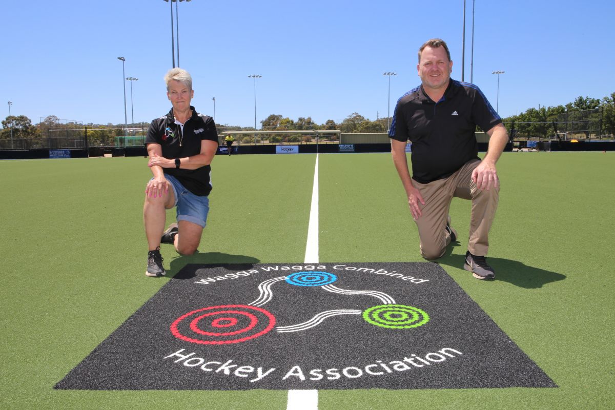 Woman and man kneeling on new synthetic surface hockey field with Wagga Wagga Combined Hockey Association insignia on surface