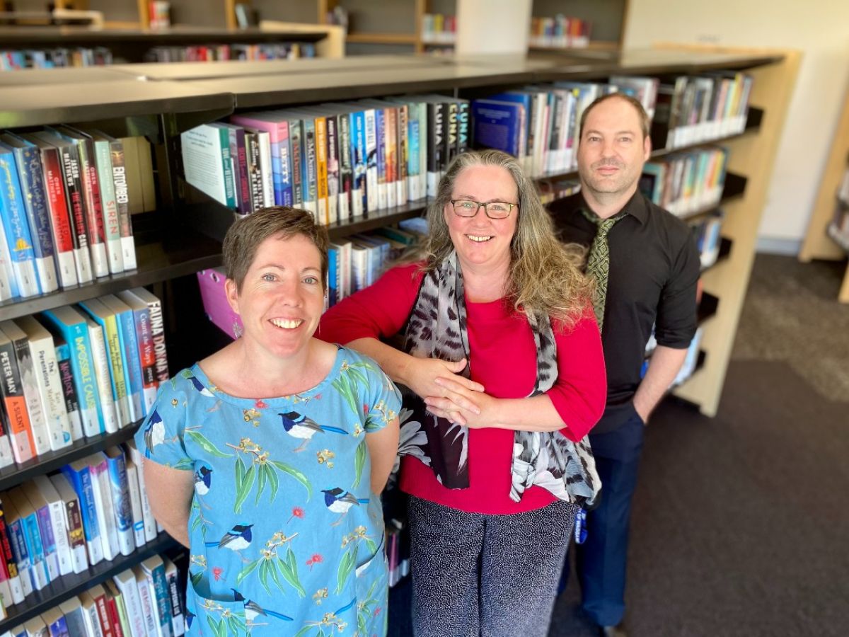 Two women and a man stand beside a bookshelf