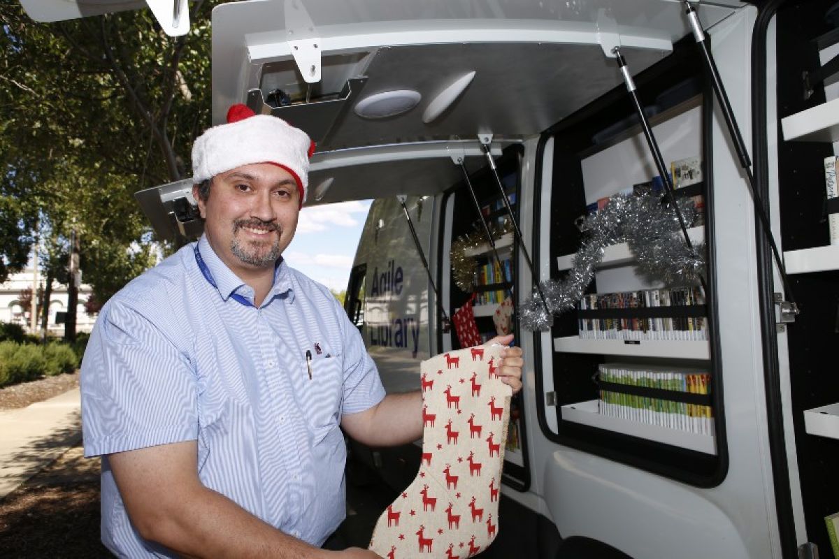 A smiling man in a Santa hat holding a Christmas stocking beside a van filled with books