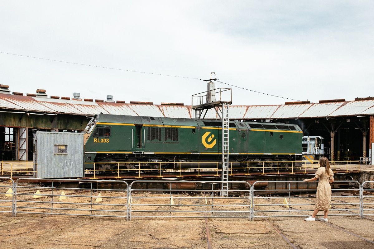 Woman in dress leaning on metal fencing barriers, looking at train engine at roundhouse in Junee