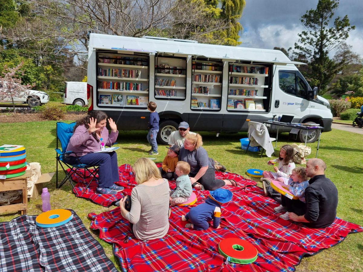 A group of children and their parents sit on a picnic rug in front of library staff member who reads them a book. The Agile Library van is on display in the background.