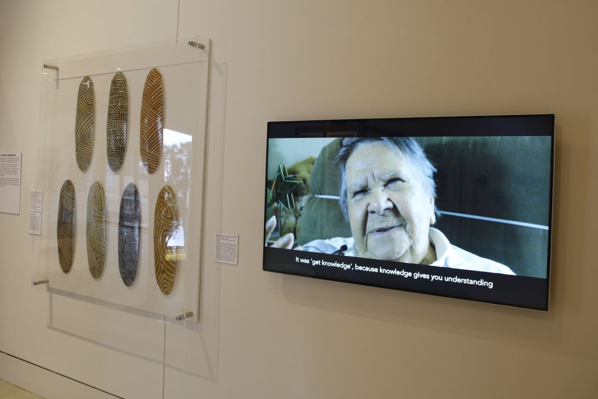 Some First Nations contemporary shields on a wall in a museum, next to screen with a woman speaking. 