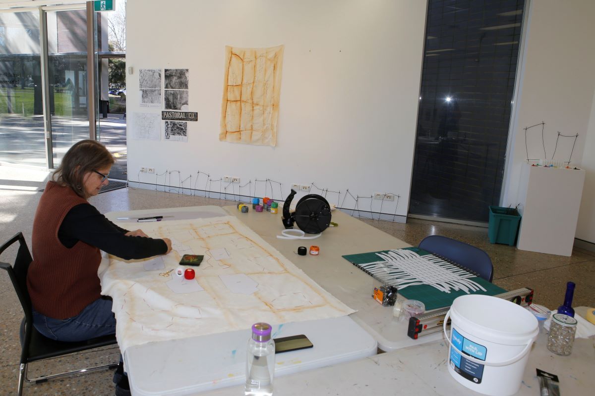 A woman sits at a table in an art space and makes artworks. 