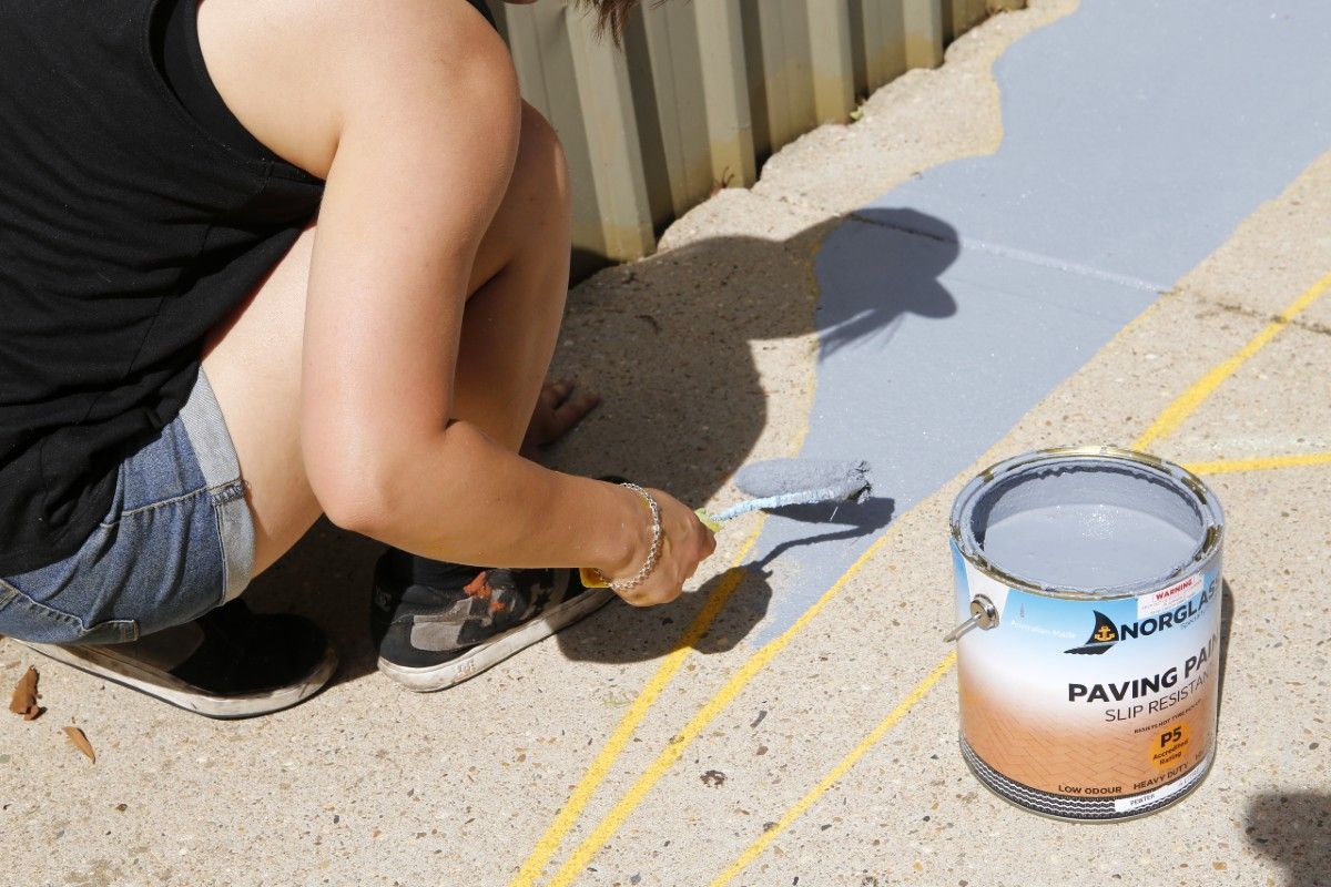 Close up shot of a students hand holding a paint roller and applying grey-blue paint to the pathway. A medium sized tin of pain sits open to the right of her hand.