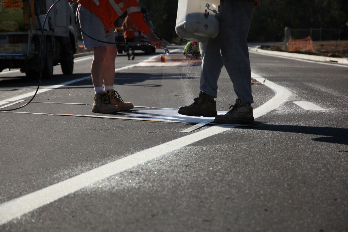 People spray white markings onto a new road surface, only the lower part of their bodies can be seen in the photo. 
