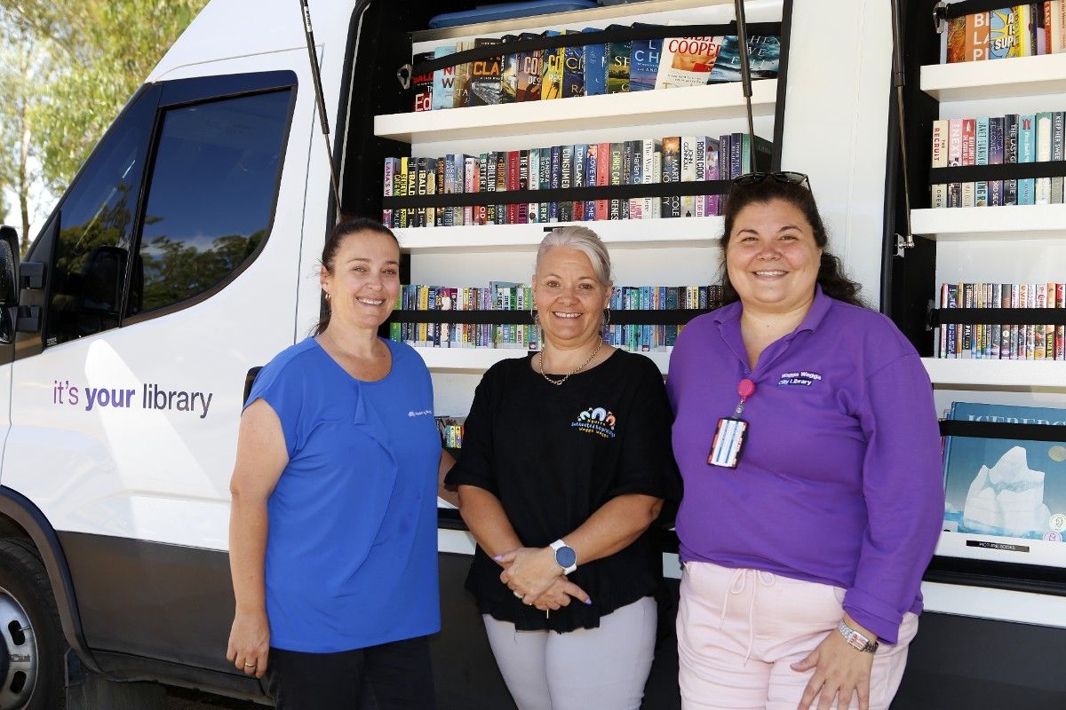 Three women standing beside Agile Library van, with books on shelving in background