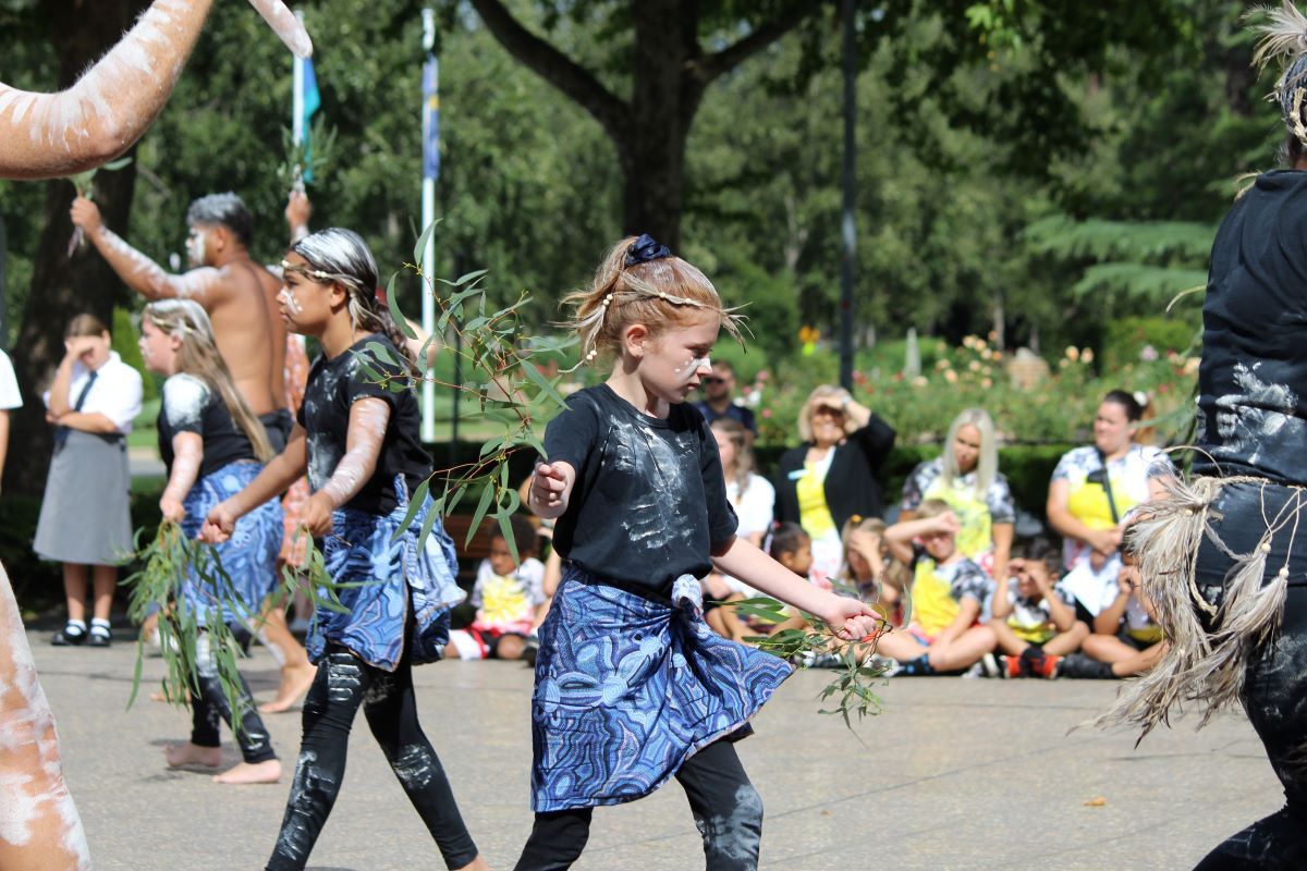 A young First Nations person dancing with a group, she is holding gum leaves and has white ochre on her face and clothing. She wears a blue skirt with an Aboriginal design. 