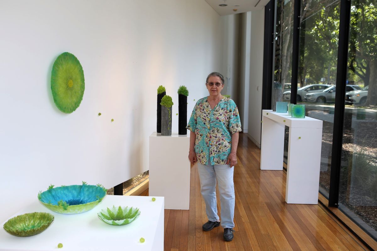 A woman stands in an art gallery space surrounded by her glass artworks. 