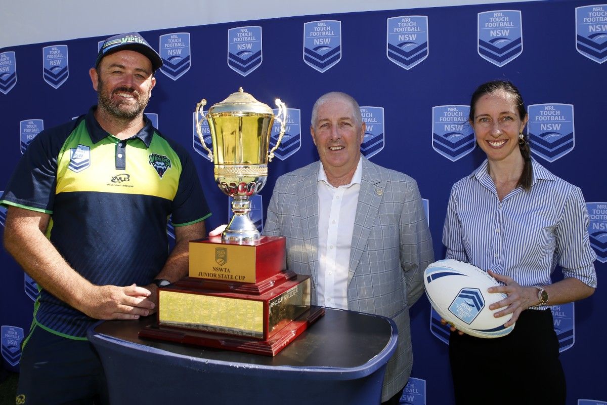 Two men and a woman standing next to a large sporting trophy, with a NSW Touch Association sign in the background.