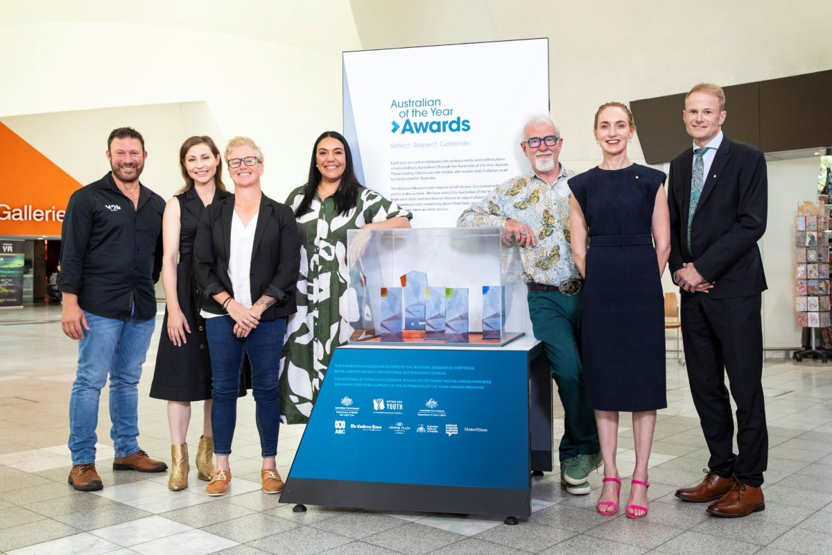 Seven adults standing in a museum setting with a sign that says Australian of the Year Awards. 