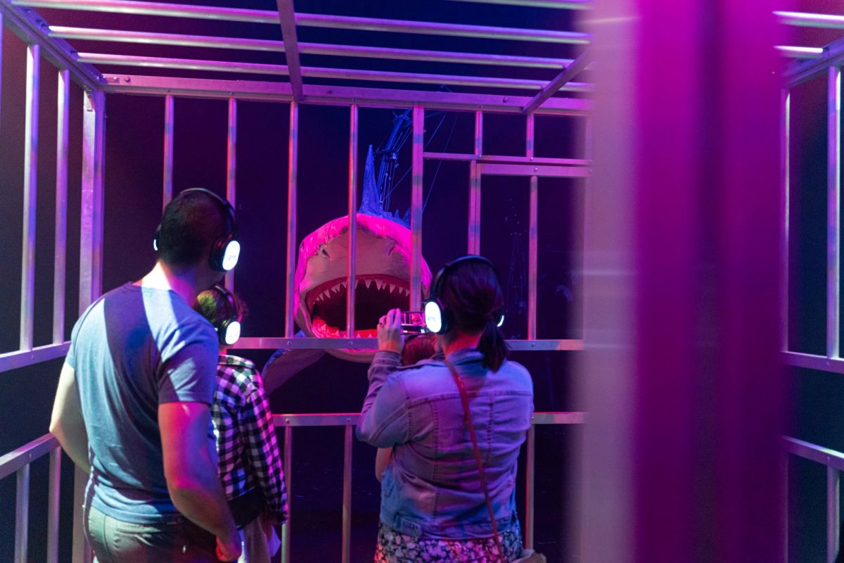 Two children stand in a large, caged exhibit resembling an underwater shark cage. The children wear headphones and are looking at a large shark puppet. 