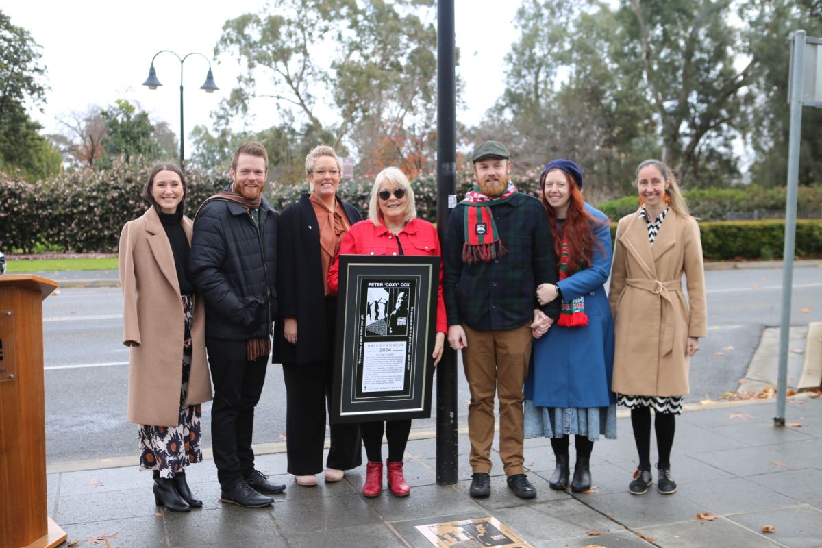 A group of people dressed in semi-formal winter clothing stand outside on a footbath lining a road. The women in the middle holds a large framed print, and on the footpath in front of them is a bronze plaque.