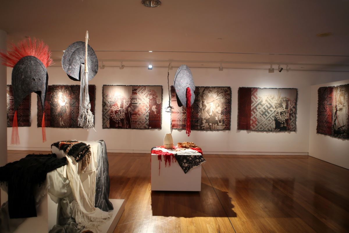 An art exhibition of textile objects hanging on the wall in the background, from the ceiling and over plinths. 