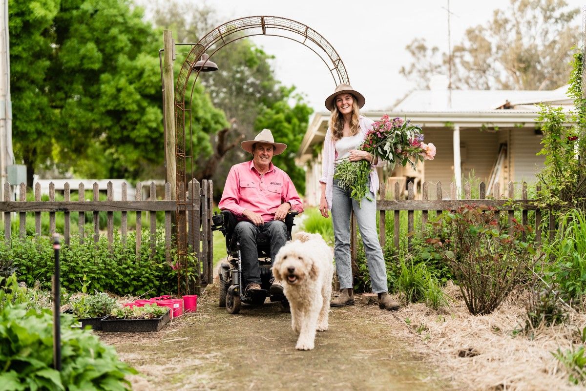 A woman holding flowers and man in a wheelchair, with their white dog, next to an archway into a garden.