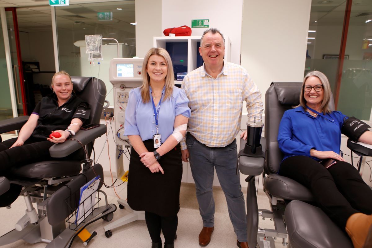 A man and woman standing between two other women who are seated in chairs and donating blood.