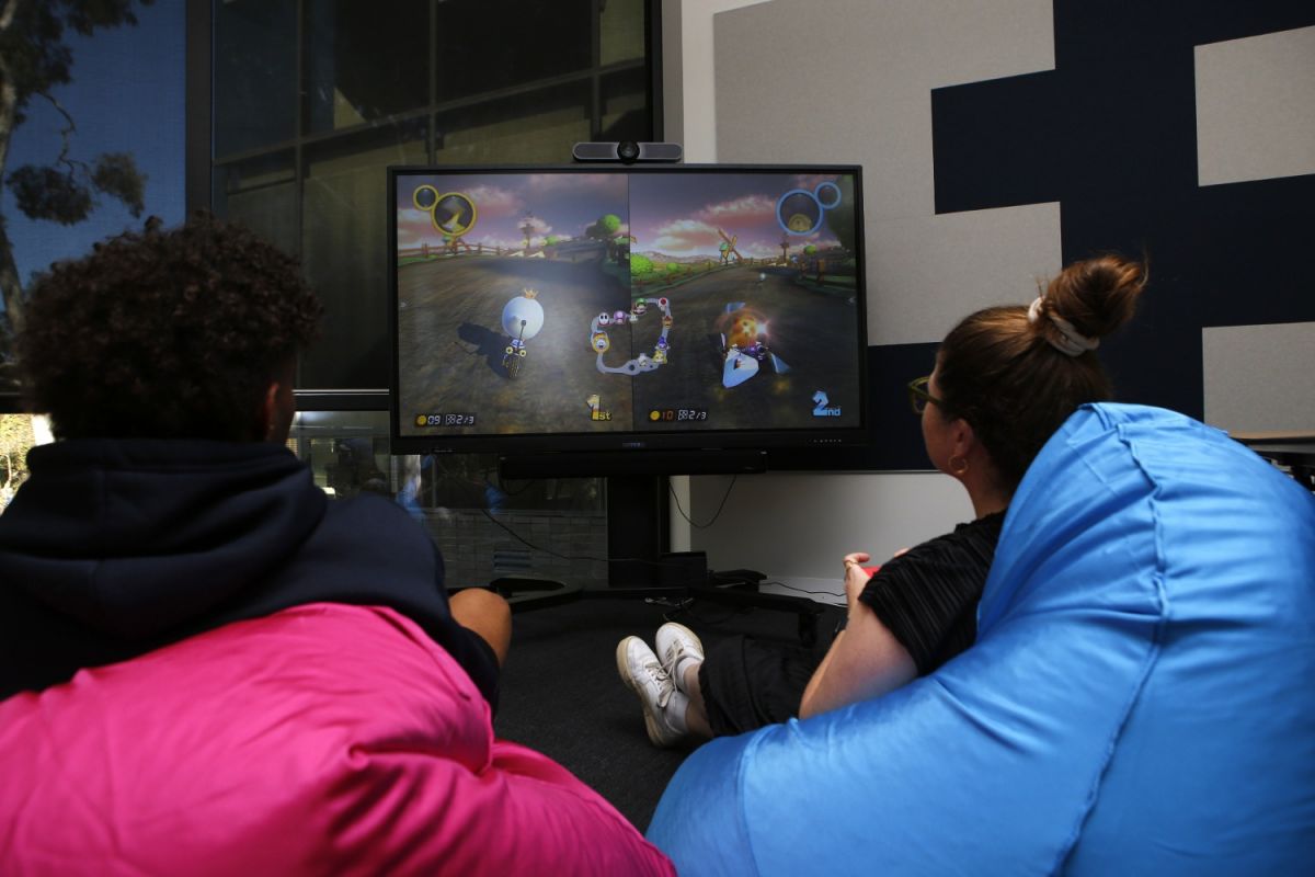 Back view of man and woman sitting on bean bags playing Mario Kart game on large TV screen in the background.