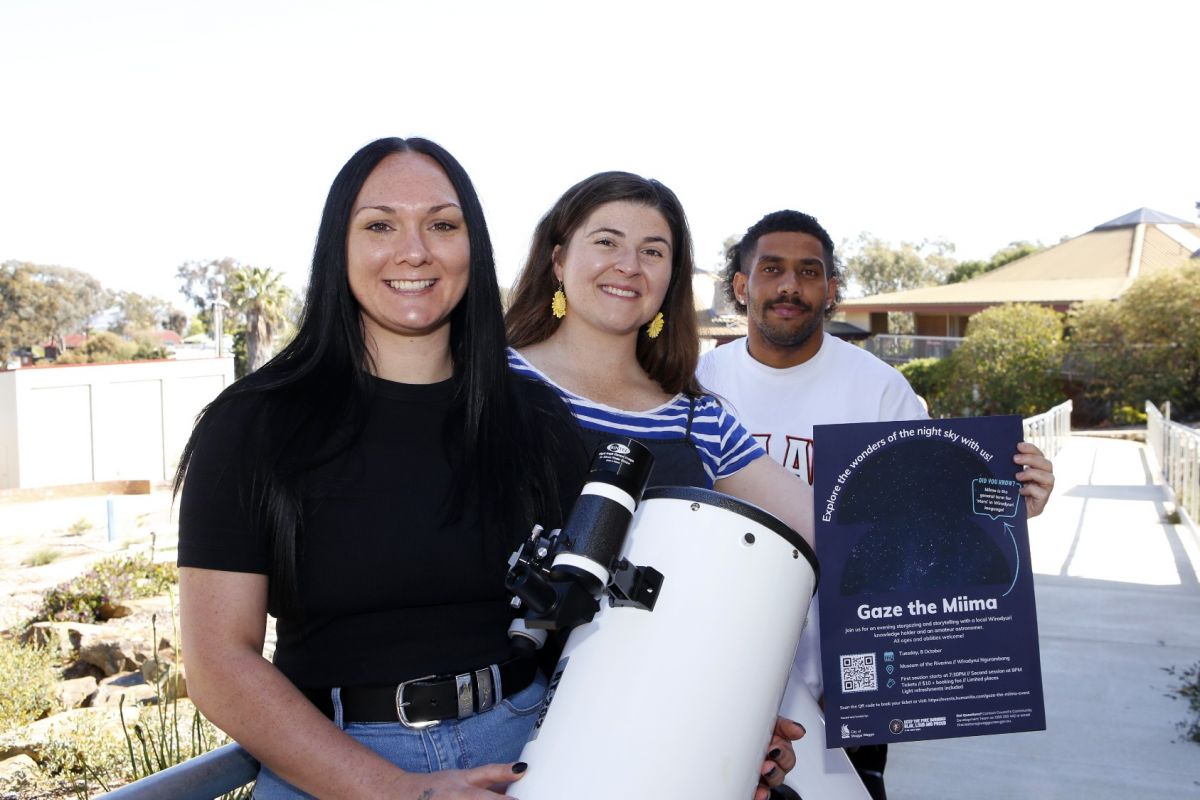 Two women and a young man standing on a verandah next to white telescopes, with one of the women holding a poster stating “Gaze the Miima”.