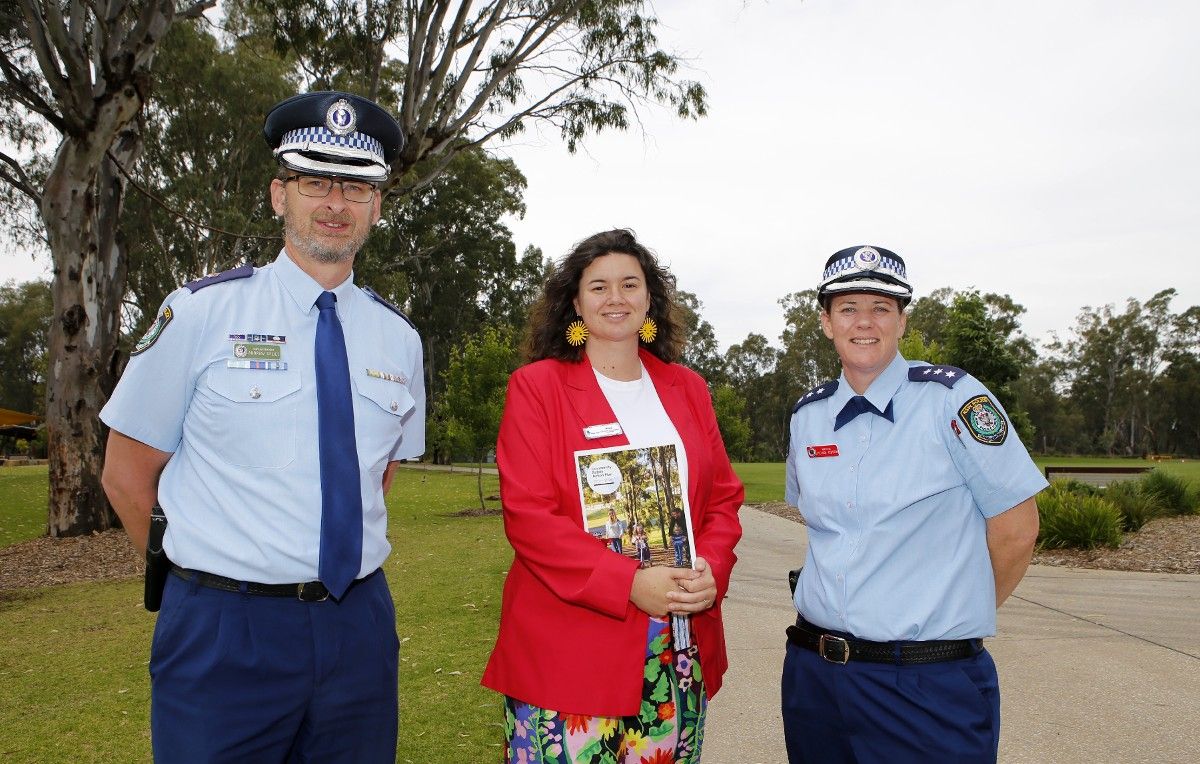 Two police officers in uniforms standing on either side of a woman wearing a red jacket and holding a copy of the Community Safety Action Plan 2022 – 2026, with all three smiling, and trees in the background.