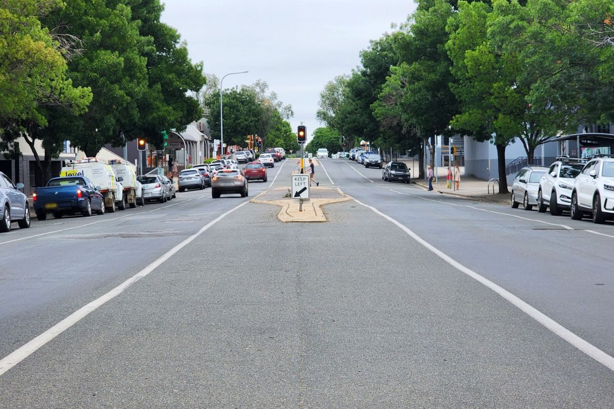 Vehicles parked on either side of a CBD road, with traffic lights at a pedestrian crossing in the background.