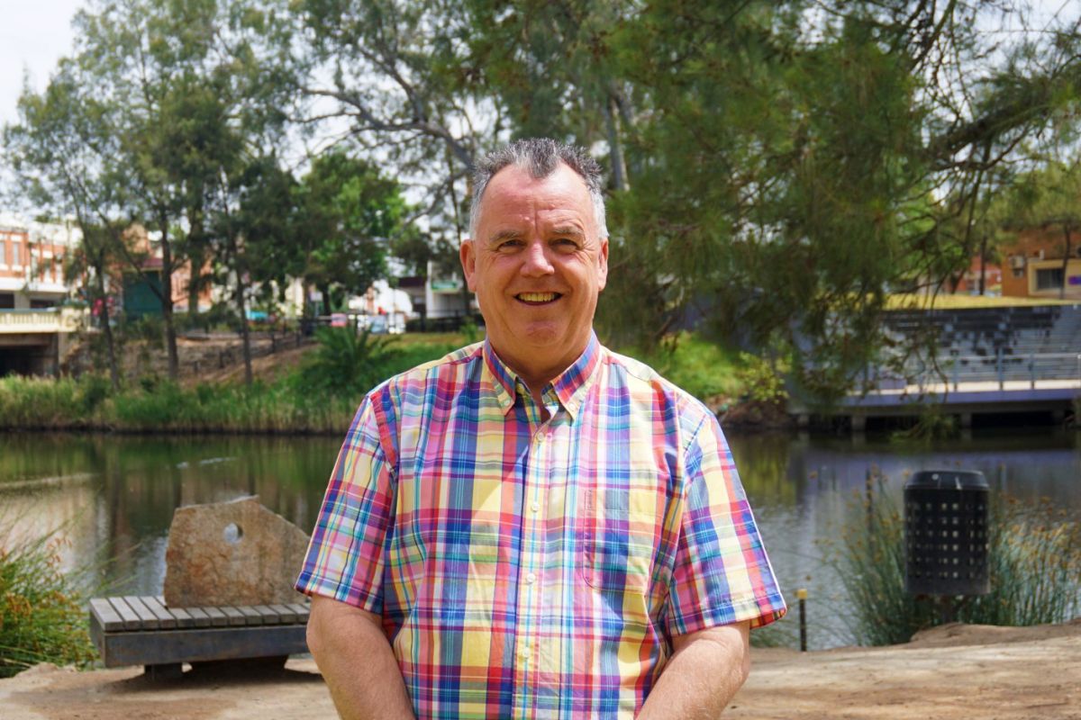 Man standing and smiling at camera, with a lagoon and outdoor amphitheatre in the background.