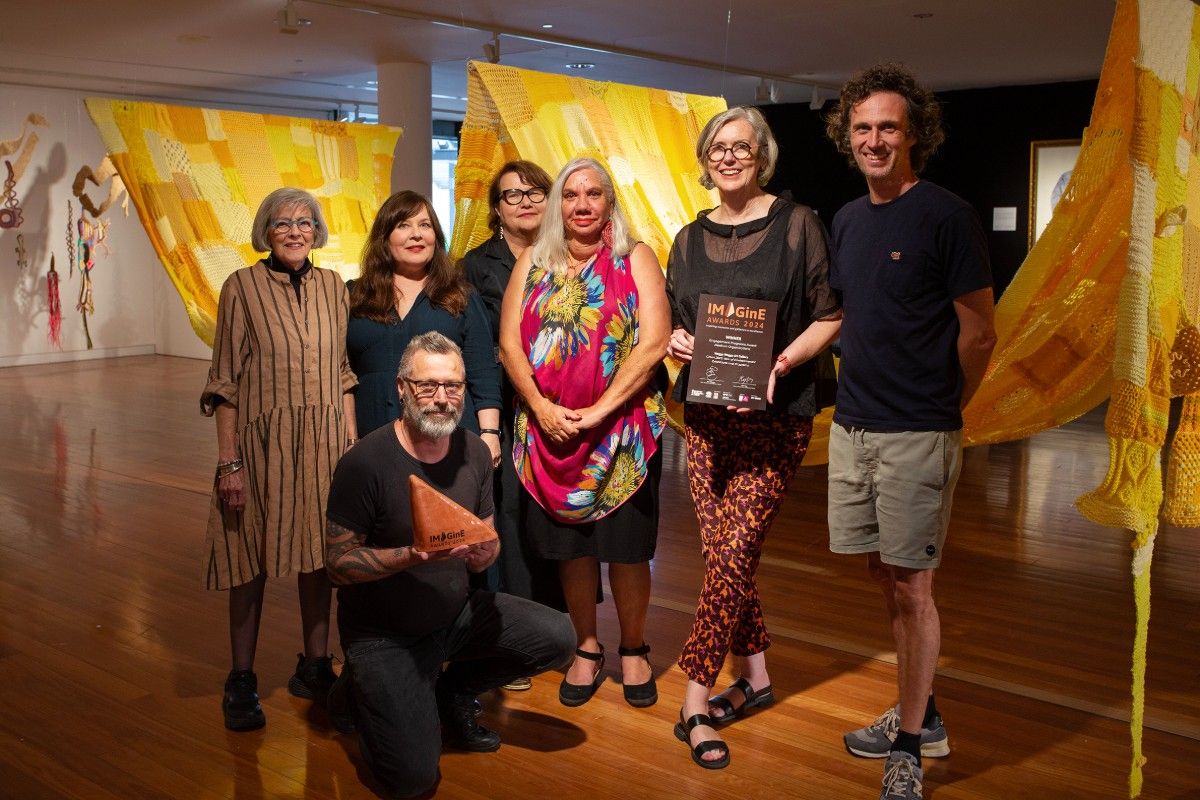 Five women and two men standing on a wooden floor in an art gallery, with exhibition pieces on the walls behind them. One of the men is holding an award.