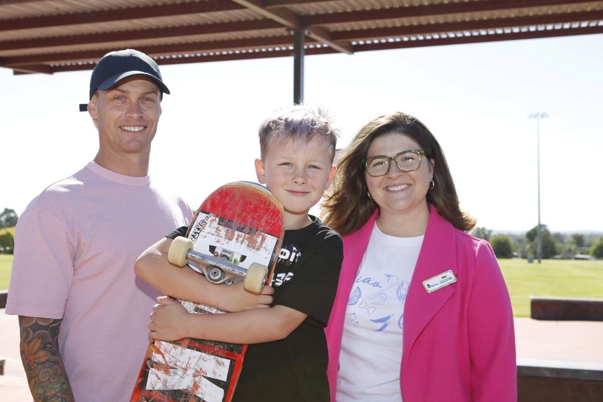 Three people stand outside. The person in the middle, a young child, holds a skateboard.