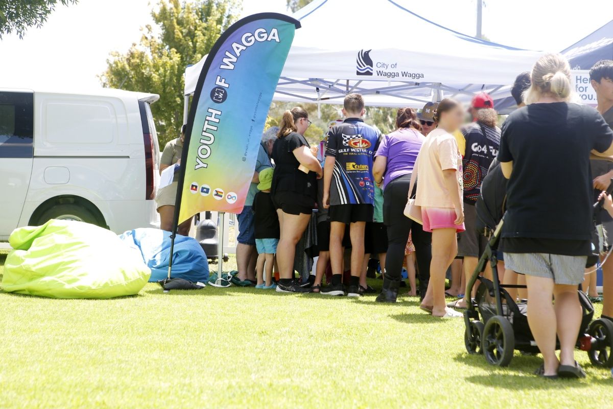 Back view of a queue of families in front of a City of Wagga Wagga marquee, with a Youth of Wagga flag next to the marquee.