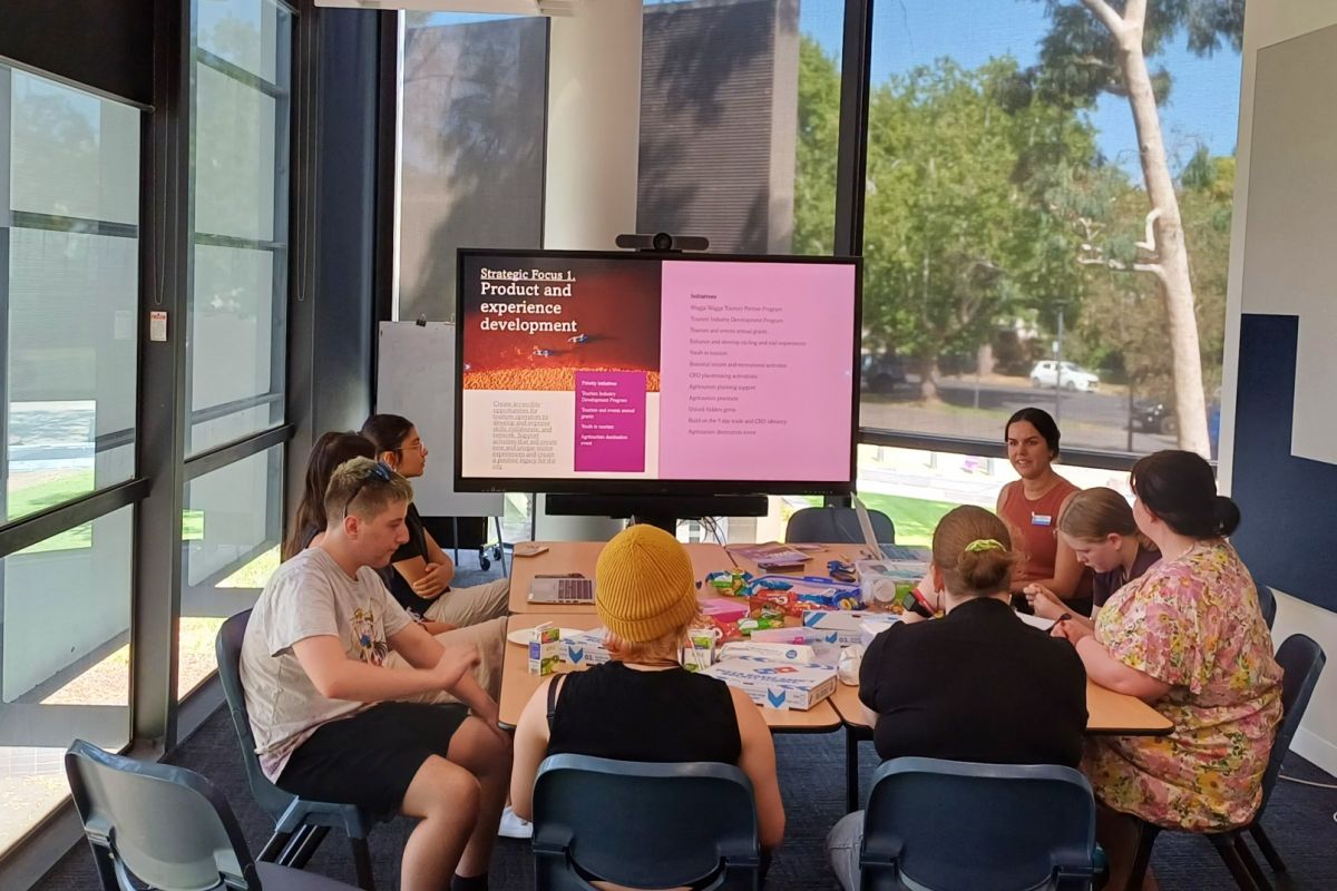 A group of young people sitting around a table covered in pizza boxes, in front of a large TV screen stating Strategic Focus 1, product and experience development.