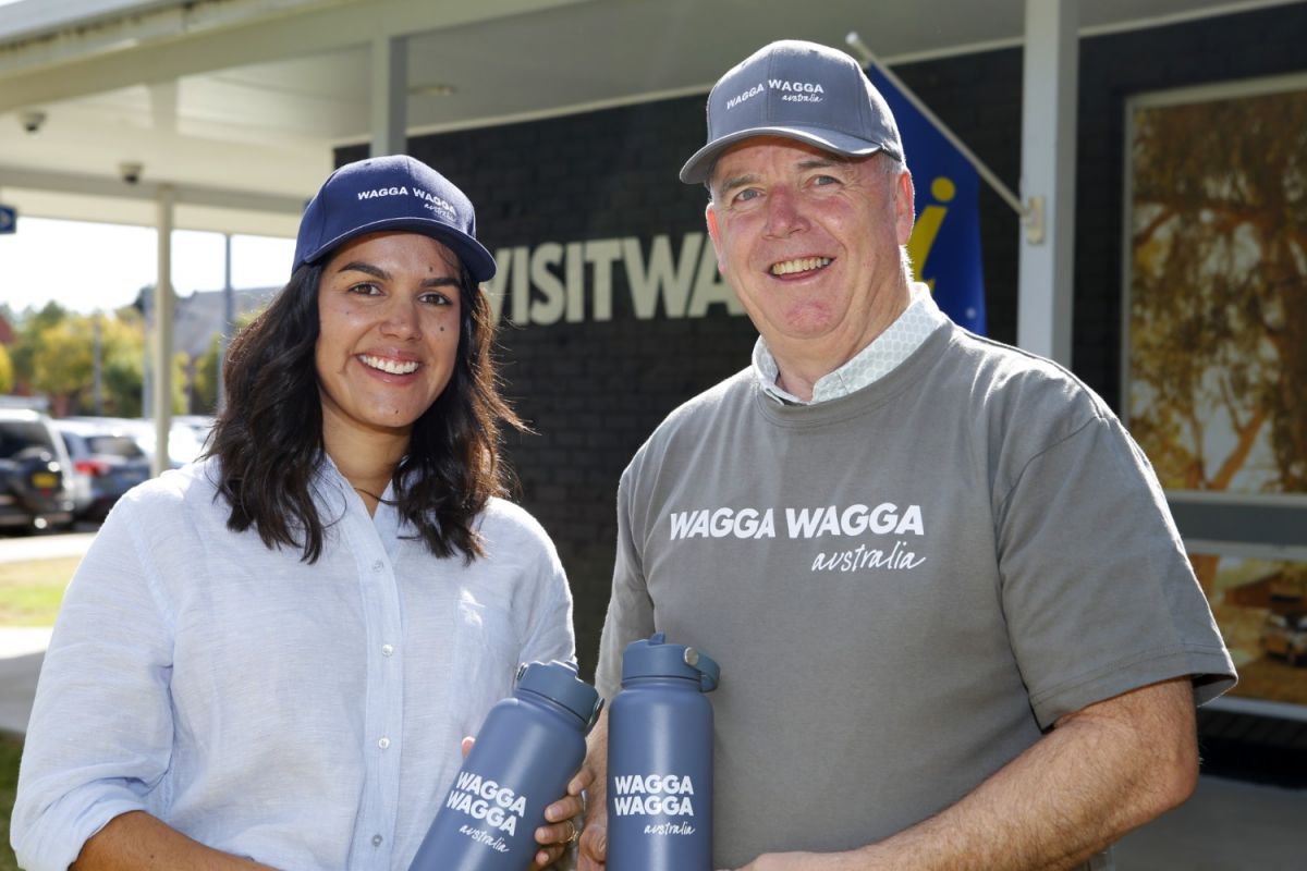 Female wearing a blue dress and a man wearing a grey tee-shirt saying Wagga Wagga Australia and both holding water bottles and wearing caps saying Wagga Wagga Australia, standing in front of a sign #visitwagga.