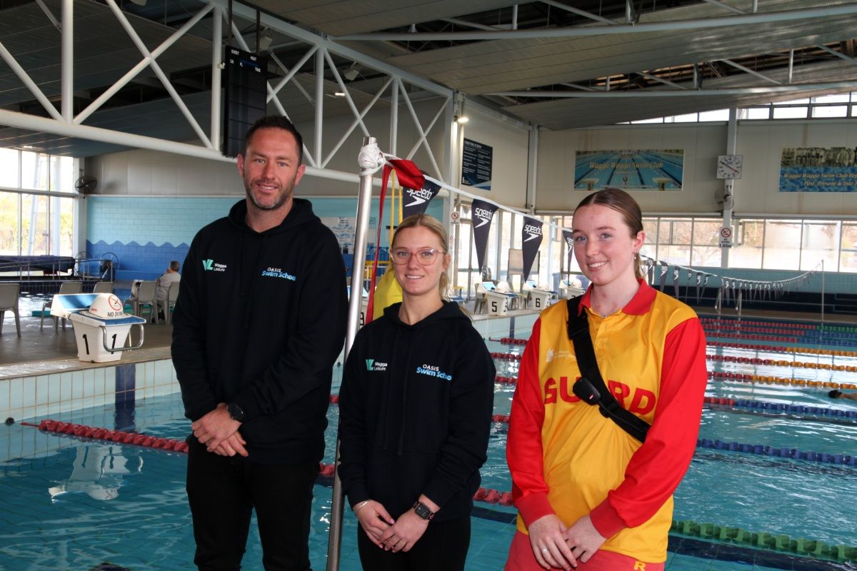 A man and a woman, both wearing black sweatshirts with the Oasis Swim School logo on the left hand side, and another female where a red and yellow life guard shirt standing beside an indoor pool.