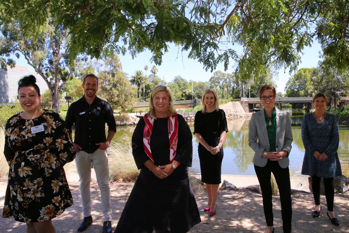 6 women and 1 man standing on grass next to lagoon