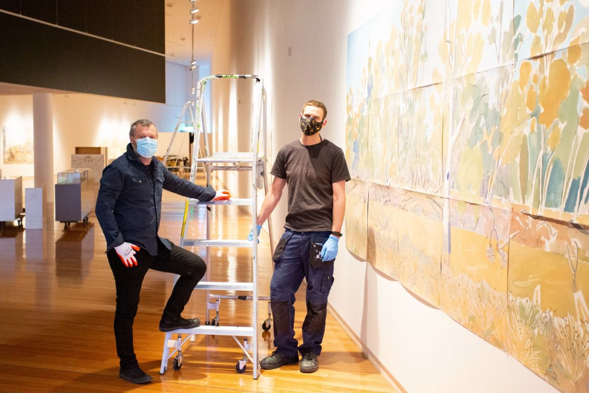 Two men standing next to ladder with large art work on wall beside them