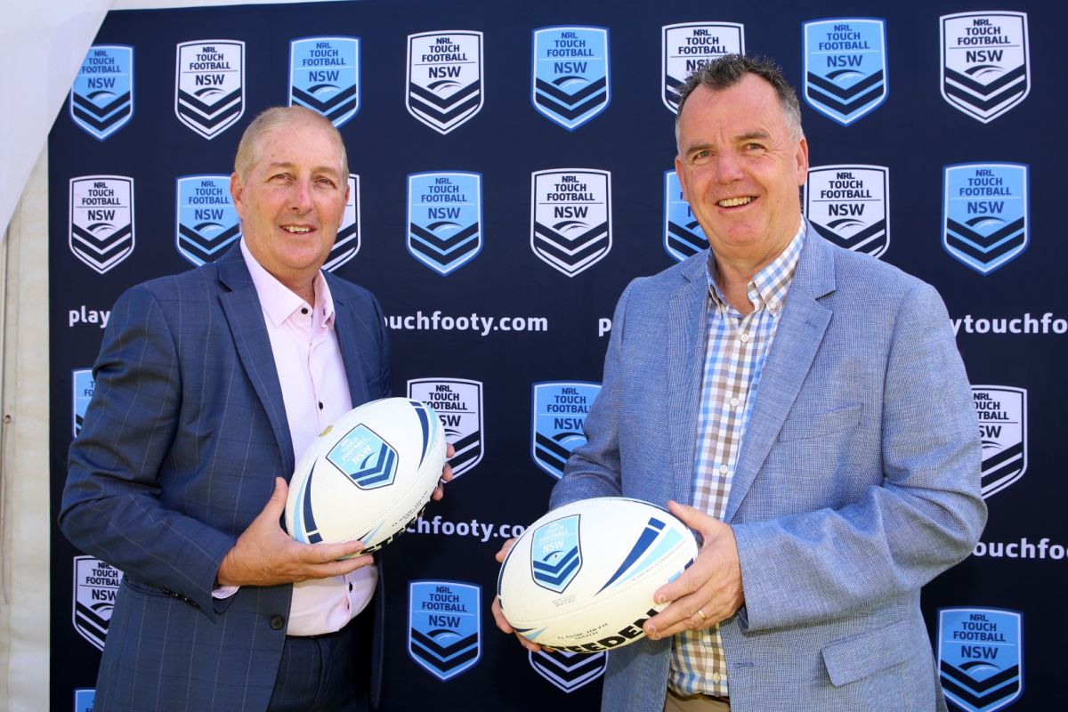 Two men holding footballs standing in front of sign with NSW Touch Association branding