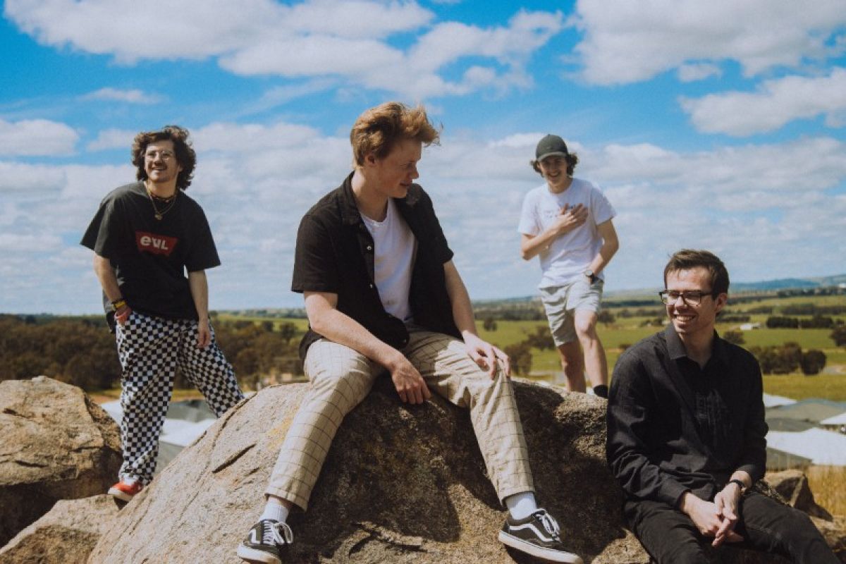 Four young men sit on a rocky outcrop in front of a blue cloudy sky