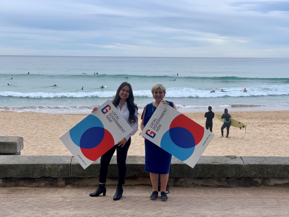 Two women in business attire, smiling and holding Local Government NSW signs, standing on paved area with beach in background