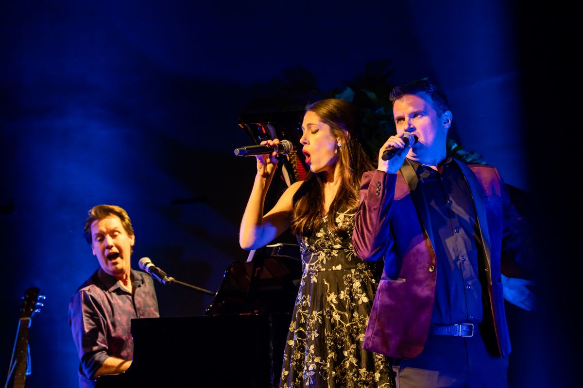 Peter J Casey, Meredith Adams, and Jamie Way on stage performing. They all sing into microphones and Mr Casey is playing the piano.