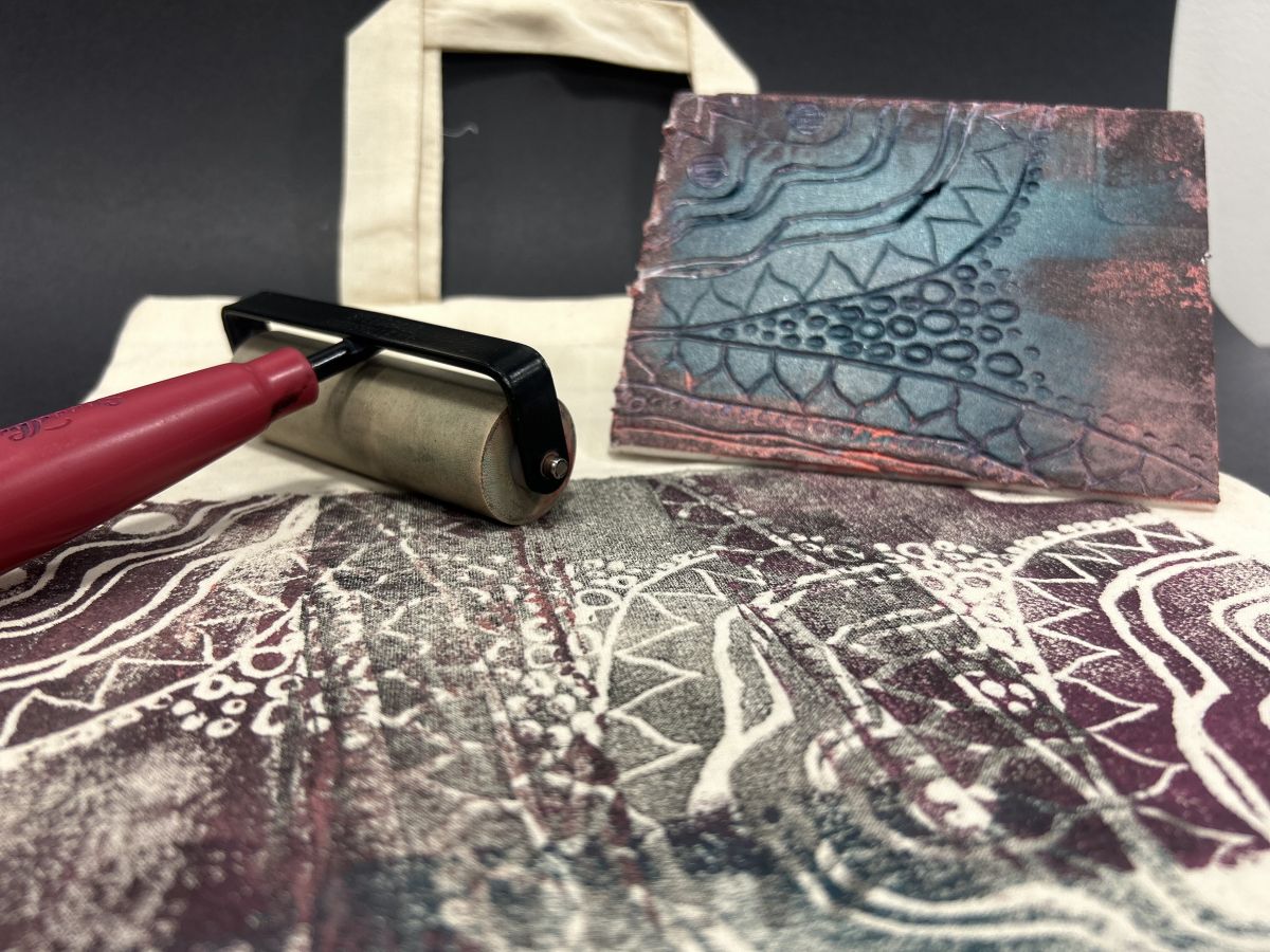 Close up of a library tote bag with fresh paint applied with lino print and roller. The lino tile and paint roller sit on the tote. 