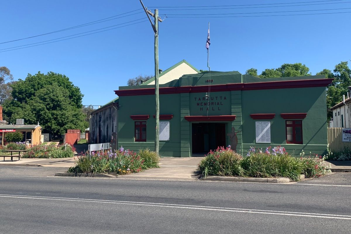 The view of a community hall from across the road on a sunny day. The building's facade is green with maroon trim. 