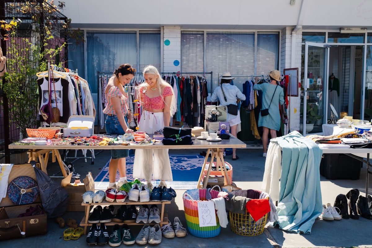 Wide shot of a garage sale held in a suburban front yard. There are two large tables with miscellaneous items and shoes at the front and three racks of clothes at the back. Two women stand at the front table looking simultaneously at an item. Another two women can be seen in the background sorting through clothes on the racks.