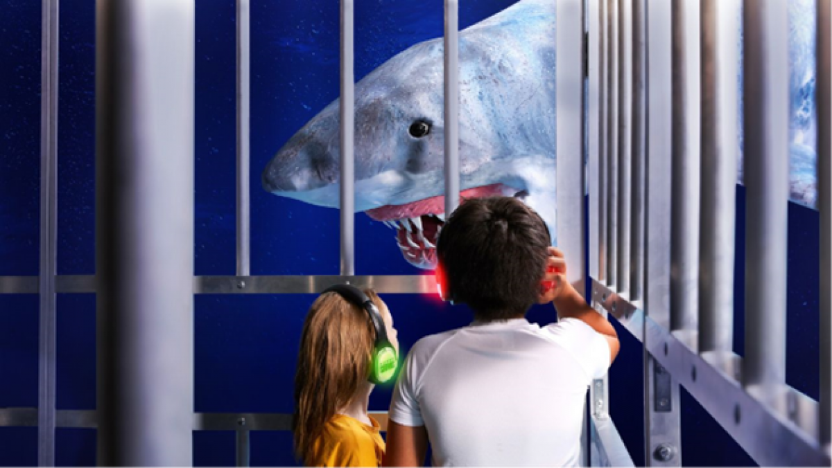 A group of people stand in a large, caged exhibit resembling an underwater shark cage. They all wear headphones and are looking at a large shark puppet.    