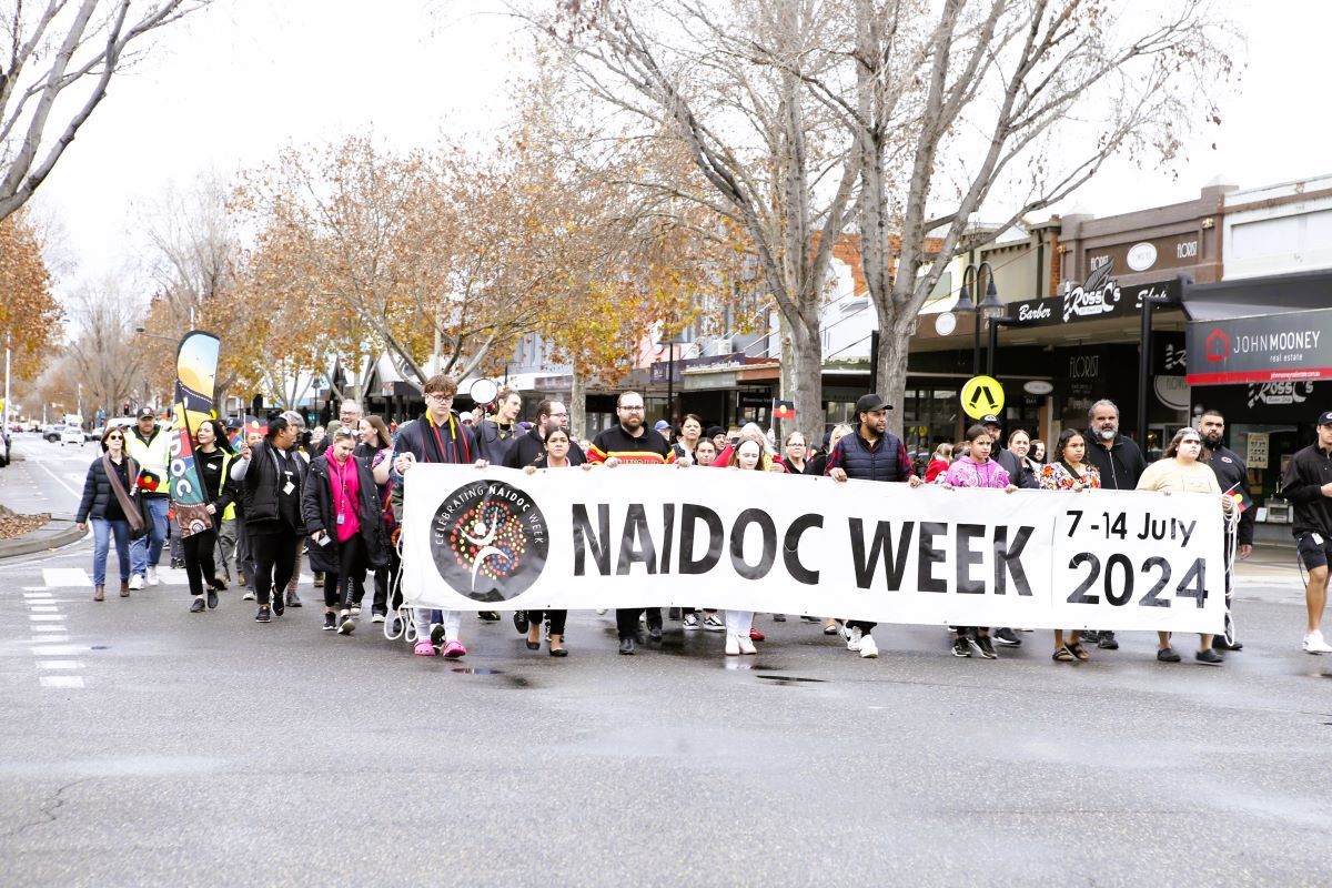 A large group of people marching down the main street of Wagga Wagga. At the front of the group people are holding a large banner that says “NAIDOC Week, 7 – 14 July 2024”.