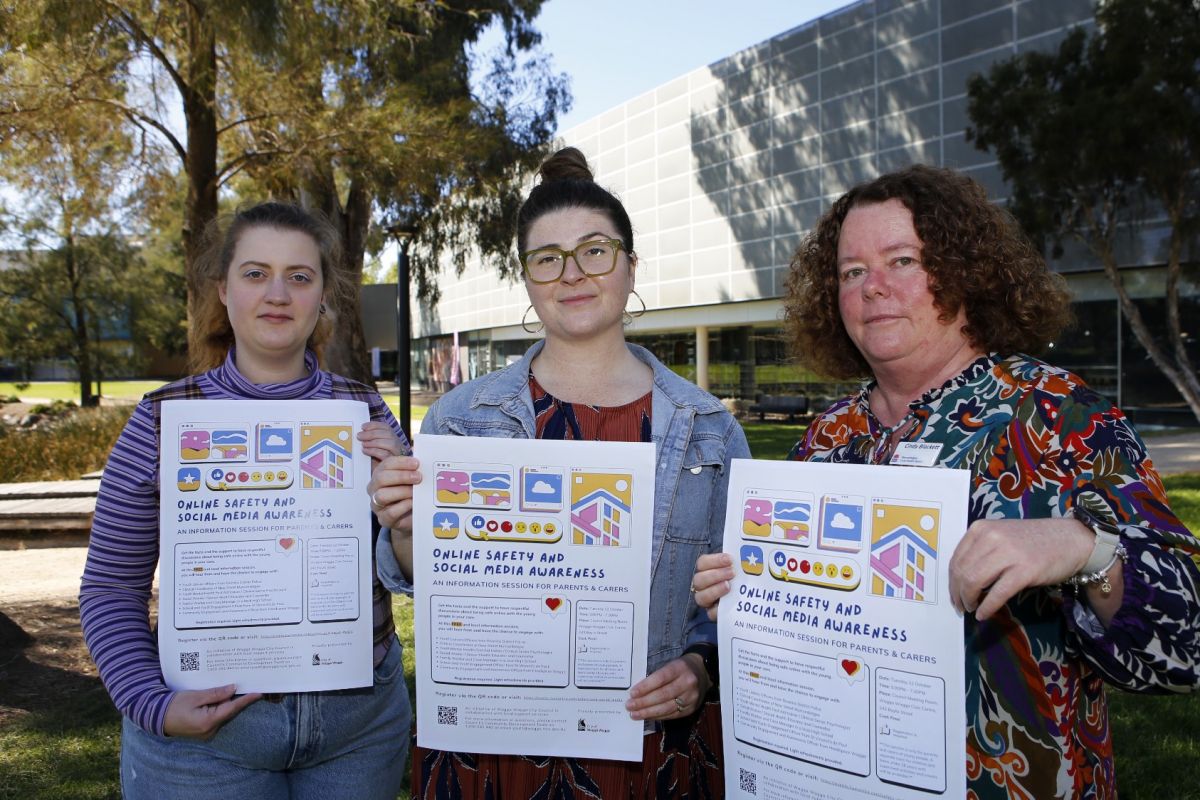 Three women standing in shade, holding posters for an Online Safety and Social Media Awareness session, with Wagga City Council Civic Centre in the background.