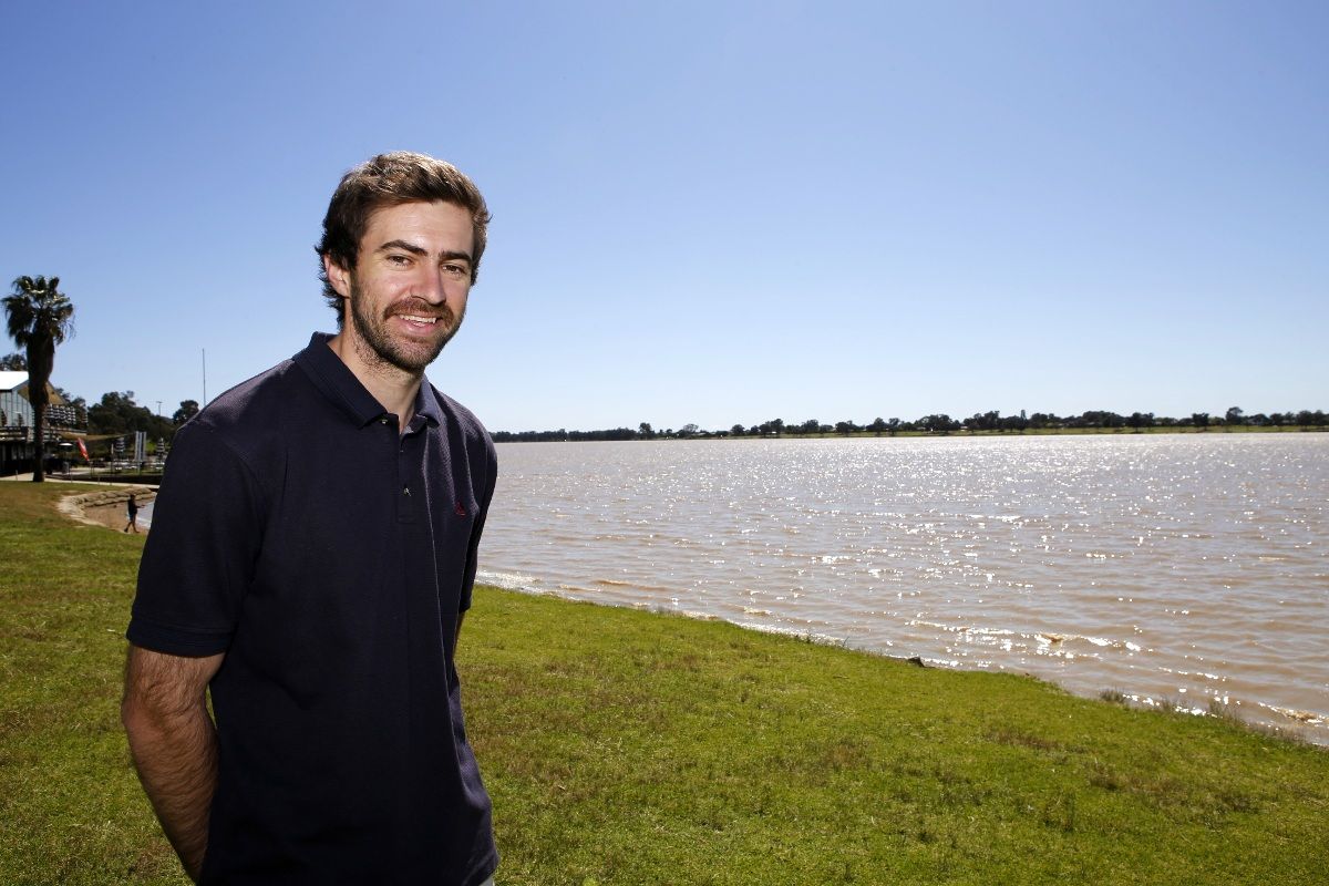 Man wearing a blue shirt, standing on grassy bank of lake. 